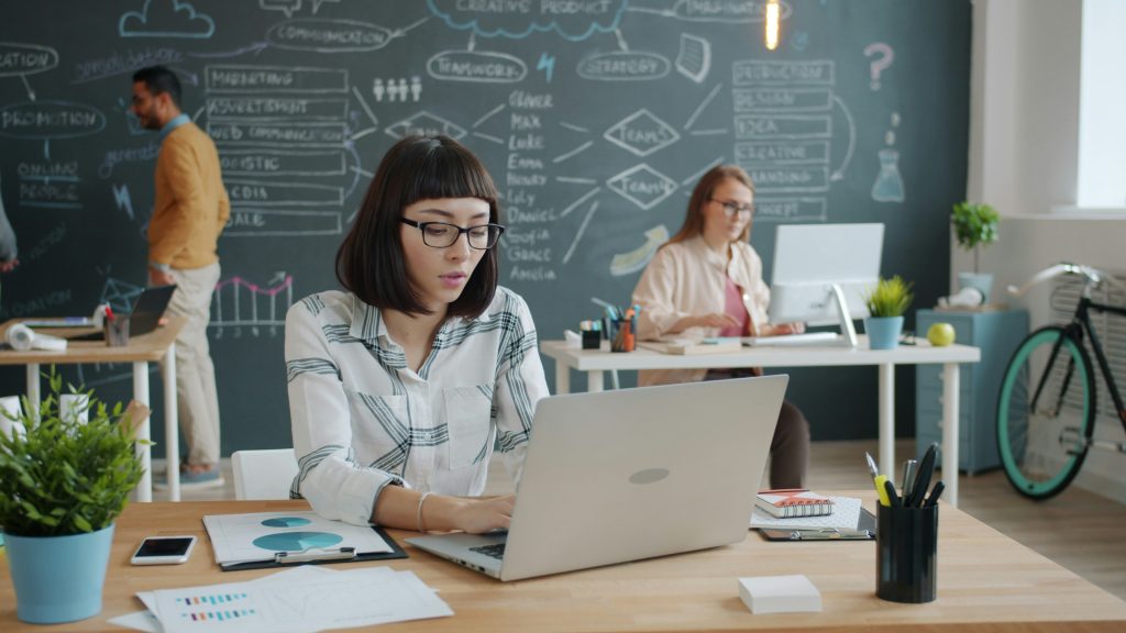 A modern office scene with several people collaborating at desks. A large chalkboard wall in the background features diagrams and notes, suggesting brainstorming or project planning in a creative workspace.