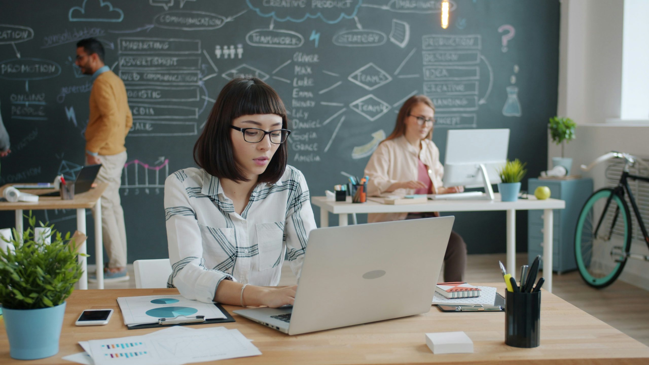 A modern office scene with several people collaborating at desks. A large chalkboard wall in the background features diagrams and notes, suggesting brainstorming or project planning in a creative workspace.